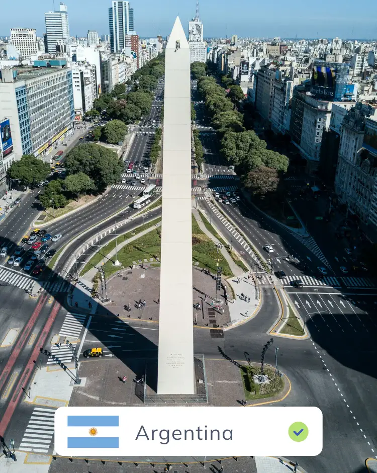 A view of the Obelisco de Buenos Aires in Argentina, a tall white monument standing amidst wide avenues and city buildings under a clear sky.