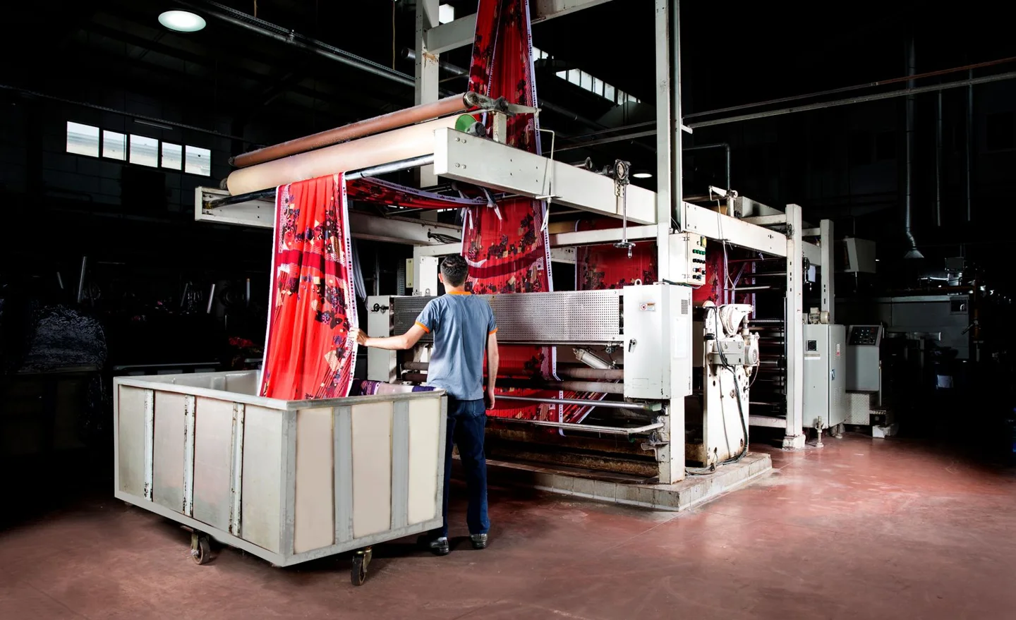 A textile worker operating a fabric printing machine on a factory floor.