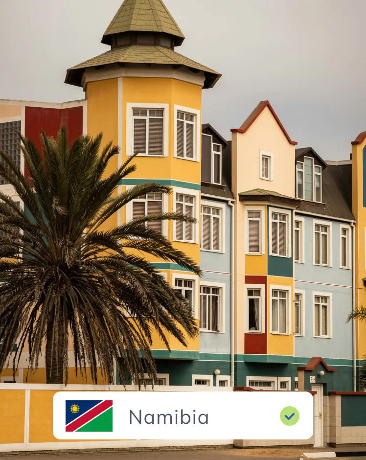 A vibrant street view in Namibia, showcasing colorful buildings with distinct architectural styles, including a prominent yellow building with a green-roofed turret, and a palm tree in the foreground.